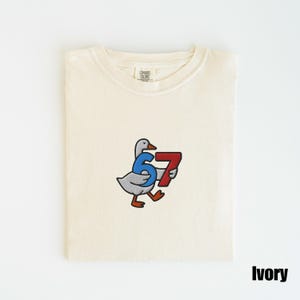 Peut inclure: T-shirt ivoire &agrave; col rond. Le t-shirt pr&eacute;sente une broderie d'une oie de dessin anim&eacute; portant le num&eacute;ro "67" en bleu et rouge. Le t-shirt est pli&eacute; proprement sur une surface blanche.