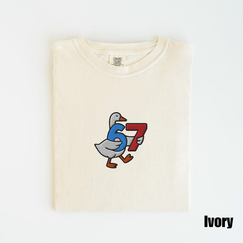 Peut inclure: T-shirt ivoire &agrave; col rond. Le t-shirt pr&eacute;sente une broderie d'une oie de dessin anim&eacute; portant le num&eacute;ro "67" en bleu et rouge. Le t-shirt est pli&eacute; proprement sur une surface blanche.