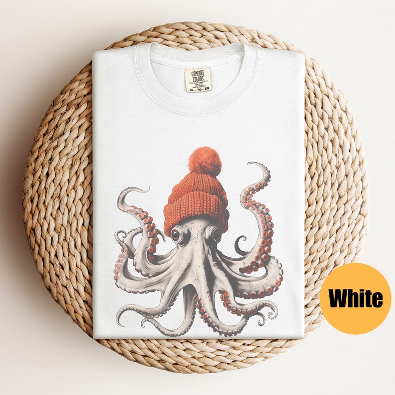 May include: White t-shirt featuring an octopus wearing an orange knit cap with a pom-pom. The octopus has detailed tentacles and is centered on the shirt. The shirt is folded on a woven, circular surface. The tag reads "Comfort Colors".