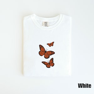 May include: White t-shirt with three embroidered orange monarch butterflies. The butterflies are in various sizes and positions, with detailed wing patterns. The shirt is folded neatly, and the word "White" is printed in a bold font at the bottom.