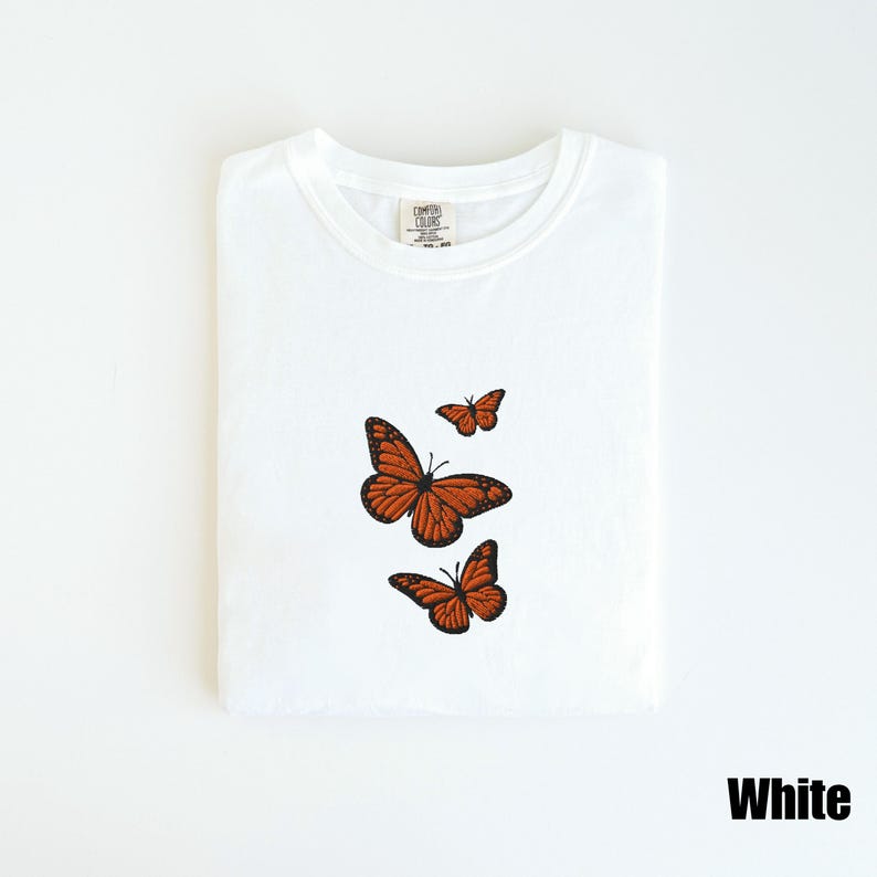May include: White t-shirt with three embroidered orange monarch butterflies. The butterflies are in various sizes and positions, with detailed wing patterns. The shirt is folded neatly, and the word "White" is printed in a bold font at the bottom.