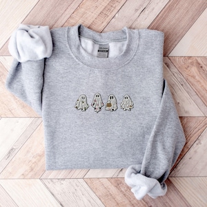 May include: A heather grey crewneck sweatshirt featuring an embroidered design of four small ghosts in a horizontal row. Each ghost has a unique pattern, including floral, celestial, and pumpkin motifs, centred on the chest of the garment.