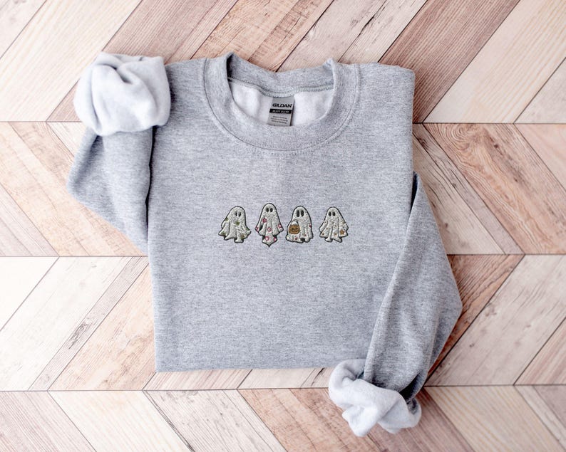 May include: A heather grey crewneck sweatshirt featuring an embroidered design of four small ghosts in a horizontal row. Each ghost has a unique pattern, including floral, celestial, and pumpkin motifs, centred on the chest of the garment.