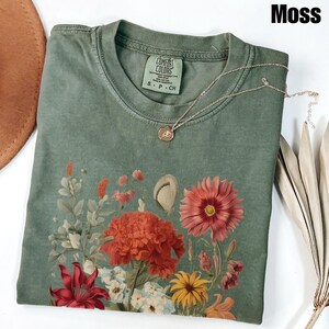 Pu&ograve; includere: Una maglietta verde con una stampa floreale che presenta una variet&agrave; di fiori in tonalit&agrave; di rosso, rosa, giallo e bianco. La maglietta ha un aspetto vissuto e il testo "COMFORT COLORS" &egrave; stampato sull'etichetta. La maglietta &egrave; etichettata "S - P - CH".