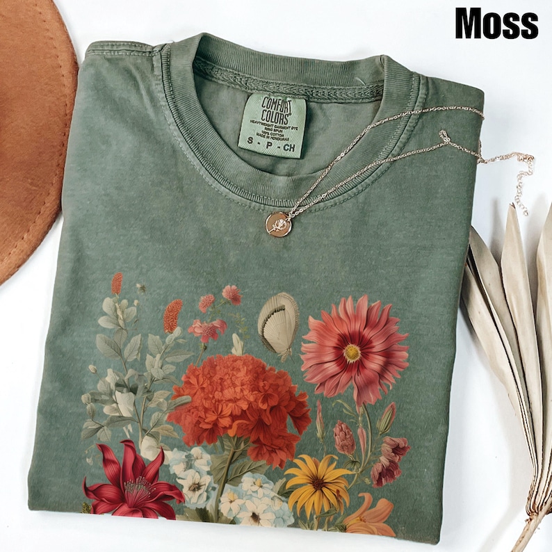 Pu&ograve; includere: Una maglietta verde con una stampa floreale che presenta una variet&agrave; di fiori in tonalit&agrave; di rosso, rosa, giallo e bianco. La maglietta ha un aspetto vissuto e il testo "COMFORT COLORS" &egrave; stampato sull'etichetta. La maglietta &egrave; etichettata "S - P - CH".