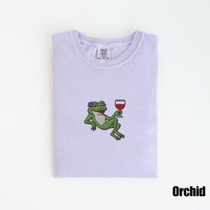 Pu&ograve; includere: T-shirt color orchidea con un ricamo a forma di rana. La rana, con un fiore e rossetto, &egrave; raffigurata mentre si rilassa tenendo in mano un bicchiere di vino rosso. La maglietta ha uno scollo tondo e un'etichetta "COMFORT COLORS". La parola "Orchid" &egrave; stampata in basso.