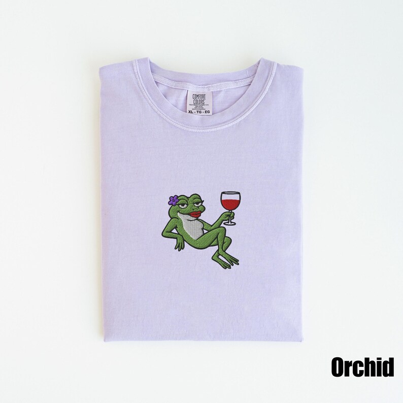 Pu&ograve; includere: T-shirt color orchidea con un ricamo a forma di rana. La rana, con un fiore e rossetto, &egrave; raffigurata mentre si rilassa tenendo in mano un bicchiere di vino rosso. La maglietta ha uno scollo tondo e un'etichetta "COMFORT COLORS". La parola "Orchid" &egrave; stampata in basso.