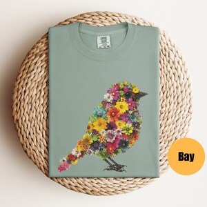 May include: A light green t-shirt with a floral bird design. The bird is made up of colorful flowers and has a light green background. The shirt has a crew neck and short sleeves.