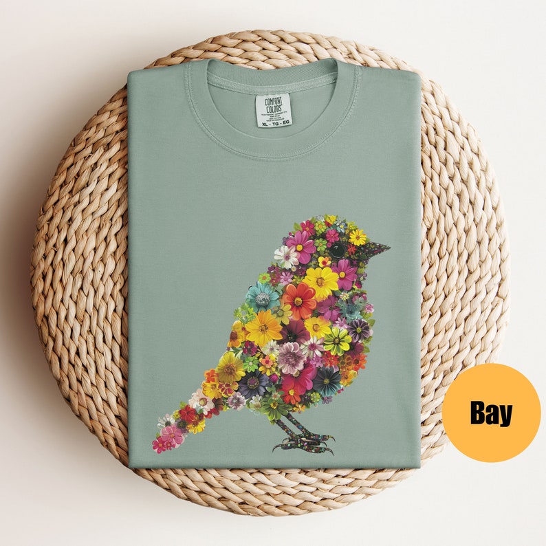May include: A light green t-shirt with a floral bird design. The bird is made up of colorful flowers and has a light green background. The shirt has a crew neck and short sleeves.
