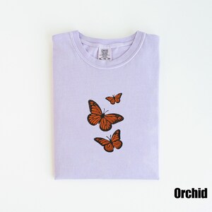 May include: A light purple t-shirt with three embroidered orange butterflies. The shirt is folded neatly and has a tag at the neckline. The word "Orchid" is printed in black at the bottom right corner.