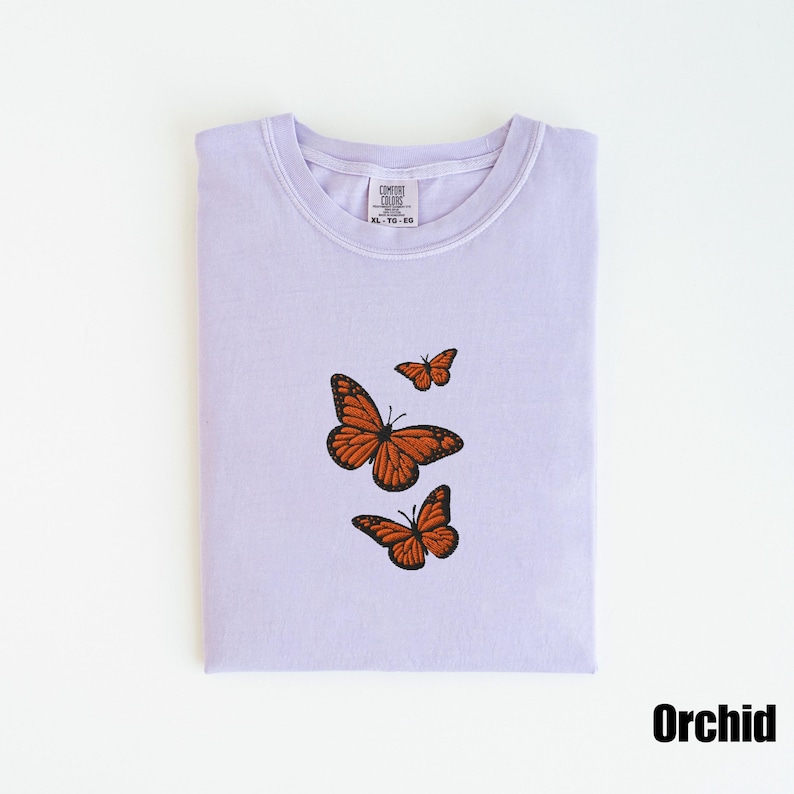 May include: A light purple t-shirt with three embroidered orange butterflies. The shirt is folded neatly and has a tag at the neckline. The word "Orchid" is printed in black at the bottom right corner.