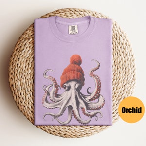May include: A lilac-colored t-shirt featuring a whimsical illustration of an octopus wearing a knitted orange beanie with a pom-pom. The octopus has detailed tentacles and large eyes. The shirt is folded on a woven, circular surface.