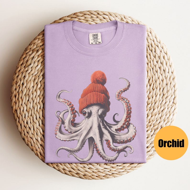 May include: A lilac-colored t-shirt featuring a whimsical illustration of an octopus wearing a knitted orange beanie with a pom-pom. The octopus has detailed tentacles and large eyes. The shirt is folded on a woven, circular surface.