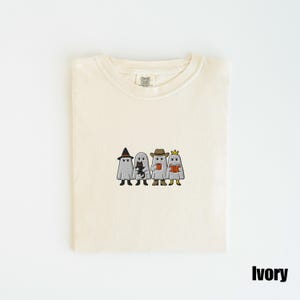 May include: Ivory-colored t-shirt with an embroidered design of four cartoon ghosts. Each ghost is dressed in a different costume: a witch hat, a cat, a cowboy hat, and a crown. The shirt is folded neatly.