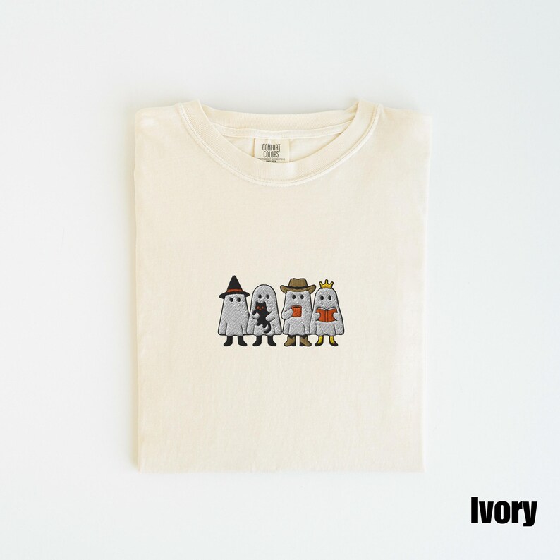 May include: Ivory-colored t-shirt with an embroidered design of four cartoon ghosts. Each ghost is dressed in a different costume: a witch hat, a cat, a cowboy hat, and a crown. The shirt is folded neatly.
