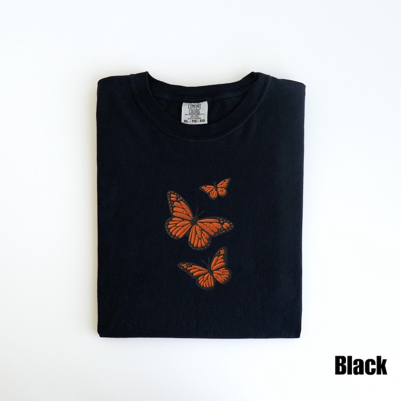 May include: Black t-shirt with an embroidered design of three orange monarch butterflies. The shirt is folded neatly and has a tag in the upper center. The word "Black" is in the lower right corner.