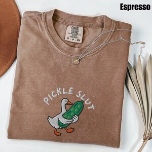May include: Espresso-colored t-shirt with the embroidered phrase "PICKLE SLUT" and a cartoon duck holding a pickle. The shirt has a crew neck and a Comfort Colors tag. A silver necklace with a small pendant is draped across the shirt.