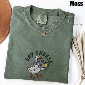 May include: A moss green t-shirt with the words "SAY CHEESE" embroidered below a cartoon goose holding a camera. A gold-colored necklace with a pendant is draped on the shirt. The shirt has a Comfort Colors tag.