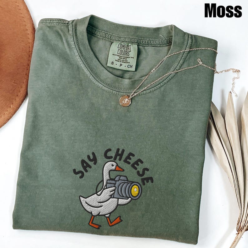 May include: A moss green t-shirt with the words "SAY CHEESE" embroidered below a cartoon goose holding a camera. A gold-colored necklace with a pendant is draped on the shirt. The shirt has a Comfort Colors tag.