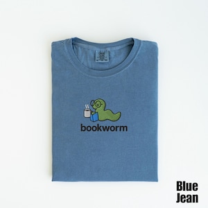 May include: Blue jean colored t-shirt with a crew neck. The shirt features an embroidered design of a green worm wearing glasses, holding a coffee cup and a book, with the word "bookworm" below. The words "Blue Jean" are in the bottom right corner.