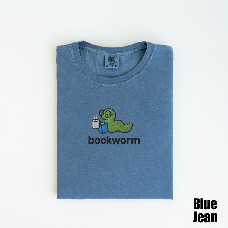 May include: Blue jean colored t-shirt with a crew neck. The shirt features an embroidered design of a green worm wearing glasses, holding a coffee cup and a book, with the word "bookworm" below. The words "Blue Jean" are in the bottom right corner.
