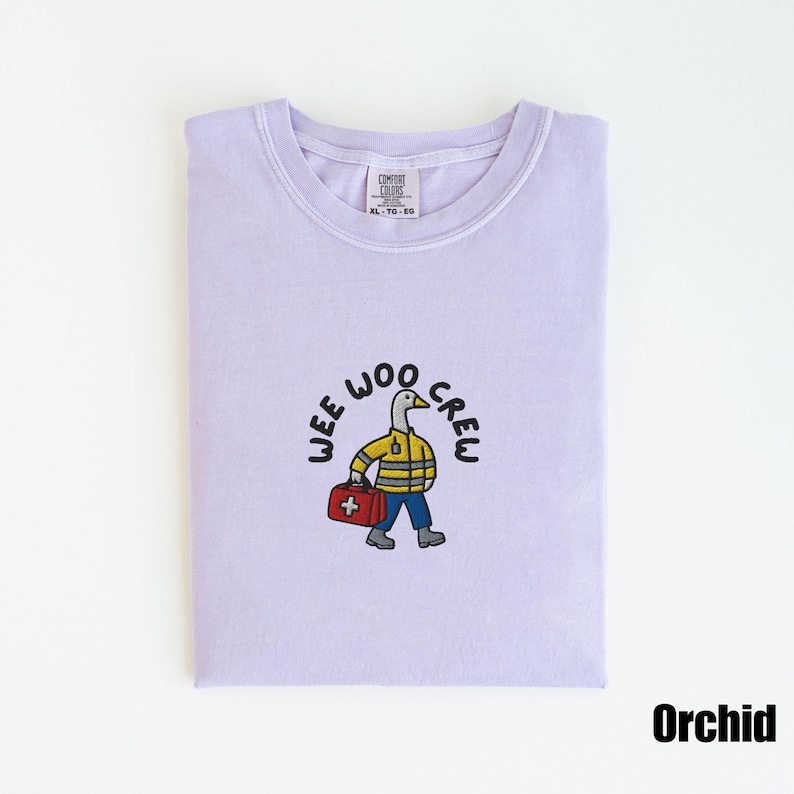 May include: A folded orchid-colored t-shirt with a crew neck. The shirt features an embroidered design of a duck wearing a yellow jacket and carrying a red medical bag, with the words "WEE WOO CREW" in a circle around the duck.