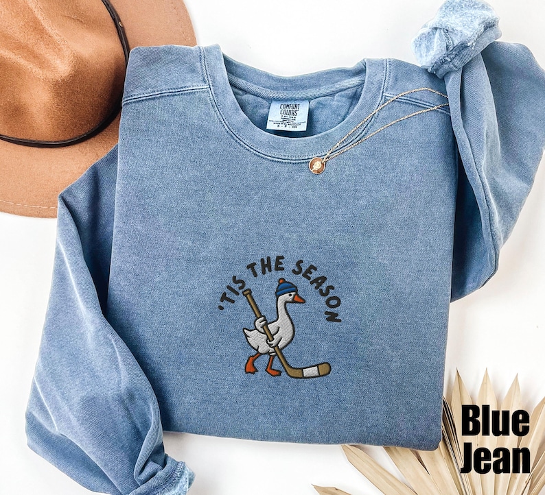 May include: Blue jean-colored sweatshirt with the phrase "'Tis the Season" embroidered above a cartoon goose wearing a hat and holding a hockey stick. The sweatshirt has a crew neck and long sleeves.