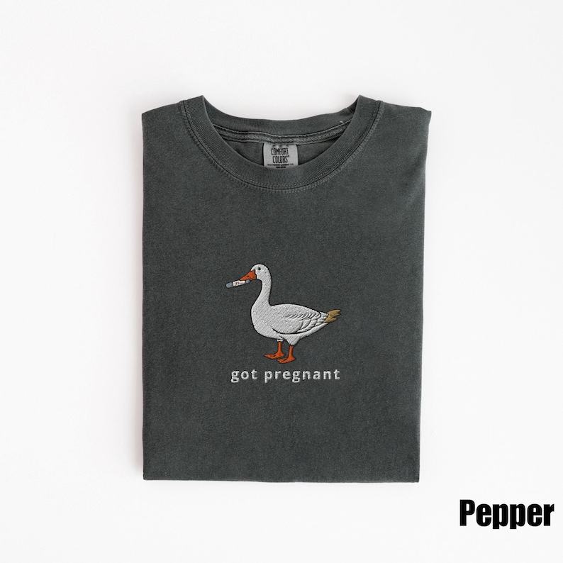 May include: Pepper-colored t-shirt with a graphic of a goose holding a thermometer. The goose is white with gray and yellow accents. The text "got pregnant" is below the goose.
