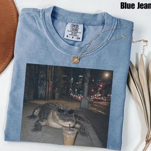 Może przedstawiać: Niebieski t-shirt w kolorze jeansu z nadrukiem aligatora w okularach przeciwsłonecznych, trzymającego mrożoną kawę na nocnej ulicy miasta. Koszulka ma okrągły dekolt i metkę marki Comfort Colors po wewnętrznej stronie kołnierza.