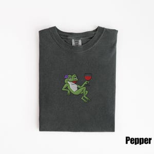 Pu&ograve; includere: T-shirt grigio scuro con un ricamo di un cartone animato a forma di rana che tiene in mano un bicchiere di vino rosso. La rana ha un fiore viola e rossetto rosso. La parola "Pepper" &egrave; stampata in bianco in basso a destra.