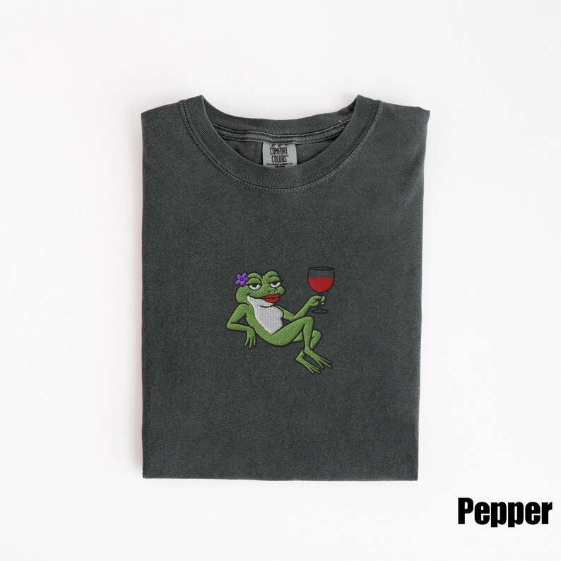 Pu&ograve; includere: T-shirt grigio scuro con un ricamo di un cartone animato a forma di rana che tiene in mano un bicchiere di vino rosso. La rana ha un fiore viola e rossetto rosso. La parola "Pepper" &egrave; stampata in bianco in basso a destra.