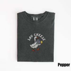 May include: A dark gray t-shirt with a crew neck. The shirt features an embroidered design of a white goose holding a camera with the words "SAY CHEESE" above it. The word "Pepper" is printed in the bottom right corner.