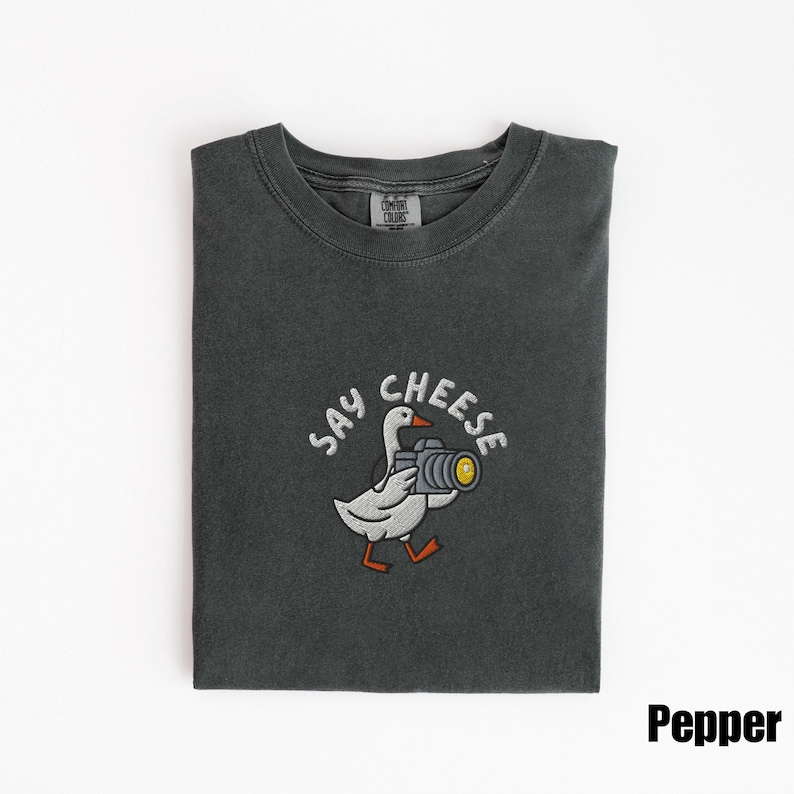 May include: A dark gray t-shirt with a crew neck. The shirt features an embroidered design of a white goose holding a camera with the words "SAY CHEESE" above it. The word "Pepper" is printed in the bottom right corner.