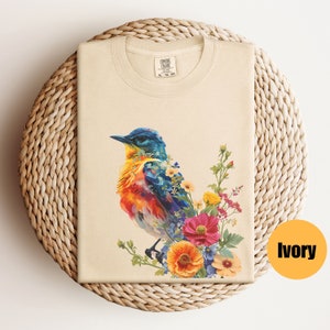 May include: A beige t-shirt featuring a colourful bird and floral design. The bird is blue, orange and yellow, and the flowers are pink, yellow and blue. The t-shirt is labelled "COMFORT COLORS" and "XL 79-80".