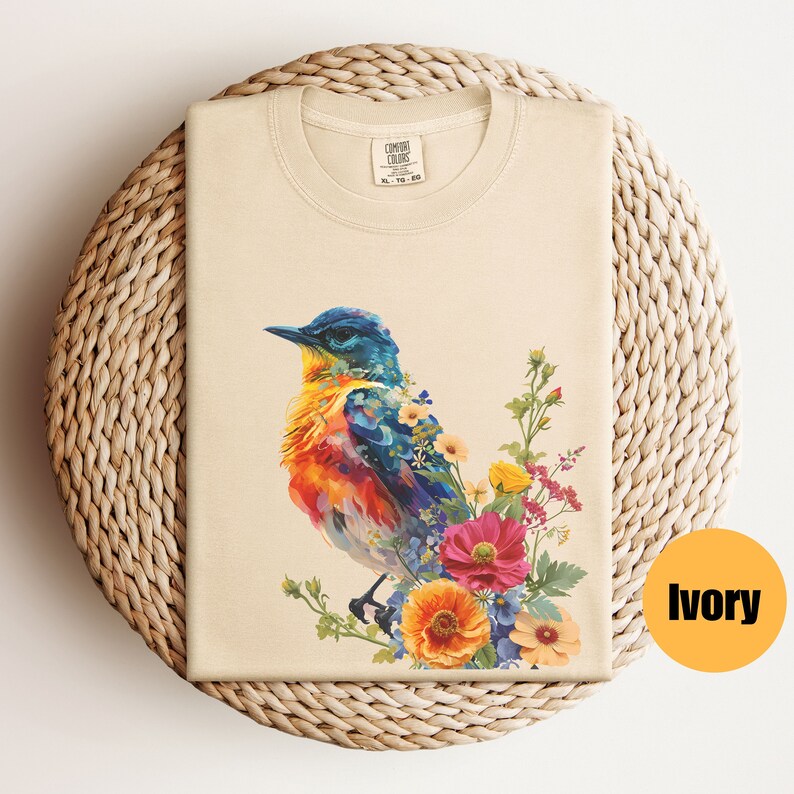 May include: A beige t-shirt featuring a colourful bird and floral design. The bird is blue, orange and yellow, and the flowers are pink, yellow and blue. The t-shirt is labelled "COMFORT COLORS" and "XL 79-80".