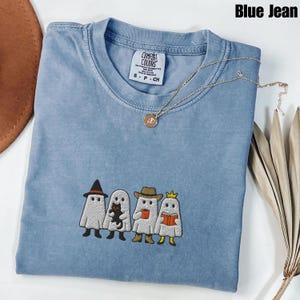 May include: Blue jean colored Comfort Colors t-shirt with an embroidered design of four ghosts, each with a unique accessory. The shirt has a crew neck and short sleeves. A gold necklace with a pendant rests on the shirt. The tag reads "Comfort Colors".