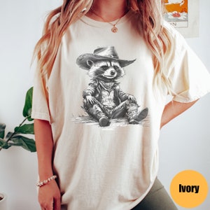 May include: A white t-shirt with a black and white illustration of a raccoon wearing a cowboy hat and leather jacket. The raccoon is sitting with its legs crossed and wearing cowboy boots. The t-shirt is labeled "ivory".