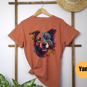 May include: A burnt orange t-shirt featuring a colourful graphic of a dog's face. The dog's fur is a mix of blue, green, purple, yellow, and orange. The dog has a black nose and brown eyes. The t-shirt has a crew neck and short sleeves.