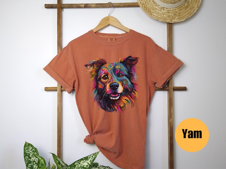 May include: A burnt orange t-shirt featuring a colourful graphic of a dog's face. The dog's fur is a mix of blue, green, purple, yellow, and orange. The dog has a black nose and brown eyes. The t-shirt has a crew neck and short sleeves.