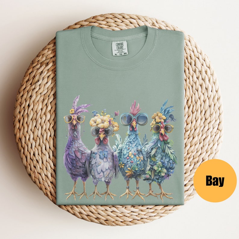 Peut inclure: Un t-shirt vert clair avec un motif de quatre poulets portant des lunettes de soleil et des fleurs. Les poulets sont violets, bleus et verts.