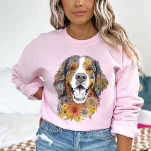 K&ouml;nnte beinhalten: Ein rosa Sweatshirt mit einem Grafikdruck eines Berner Sennenhundes mit einem Blumenmuster. Der Hund l&auml;chelt und tr&auml;gt ein Blumenhalsband.