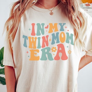 Może przedstawiać: Biały T-shirt z napisem "In My Twin Mom Era" w kolorowych, retro literach. Litery są obwiedzione czarną ramką i mają styl lat 70.