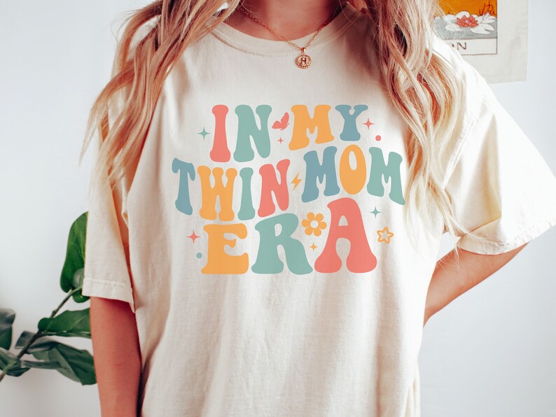 Może przedstawiać: Biały T-shirt z napisem "In My Twin Mom Era" w kolorowych, retro literach. Litery są obwiedzione czarną ramką i mają styl lat 70.