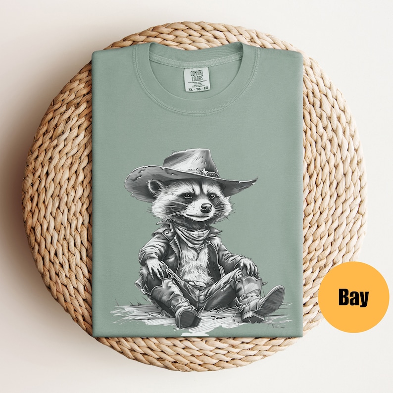 May include: A light green t-shirt with a black and white illustration of a raccoon wearing a cowboy hat and a leather jacket. The raccoon is sitting with its legs crossed and is wearing boots. The t-shirt has the text "COMFORT COLORS" printed on the tag.