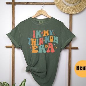 Może przedstawiać: Zielona koszulka z napisem "In My Twin Mom Era" w kolorowych, retro literach. Koszulka jest wykonana z konopi.