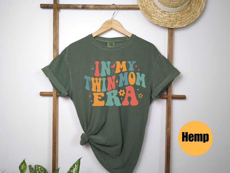 Może przedstawiać: Zielona koszulka z napisem "In My Twin Mom Era" w kolorowych, retro literach. Koszulka jest wykonana z konopi.