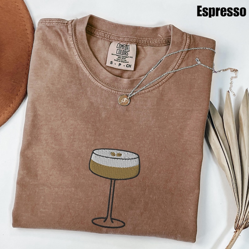 Może przedstawiać: Koszulka w kolorze espresso z grafiką kieliszka do koktajli. Koszulka ma okrągły dekolt i metkę marki Comfort Colors. Srebrny naszyjnik z małą zawieszką spoczywa na koszulce. Koszulka jest w rozmiarze S.