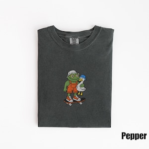 May include: Pepper-colored t-shirt featuring an embroidered graphic of a frog wearing a hat and overalls, holding a duck, and riding a skateboard. The frog has round glasses and orange sneakers. The duck wears a blue hat.