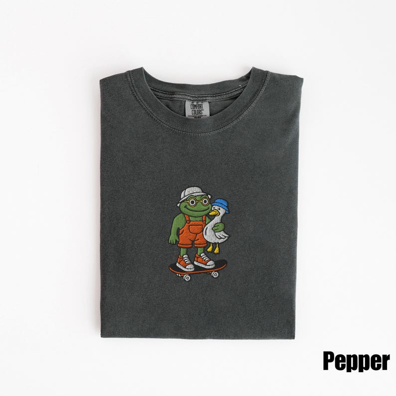 May include: Pepper-colored t-shirt featuring an embroidered graphic of a frog wearing a hat and overalls, holding a duck, and riding a skateboard. The frog has round glasses and orange sneakers. The duck wears a blue hat.