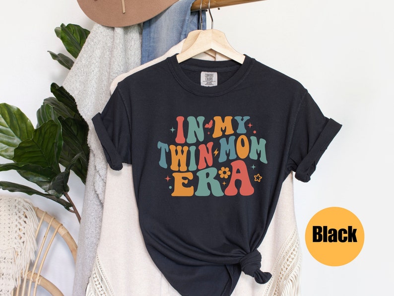 Może przedstawiać: Czarny T-shirt z napisem "In My Twin Mom Era" w kolorowych, retro literach.